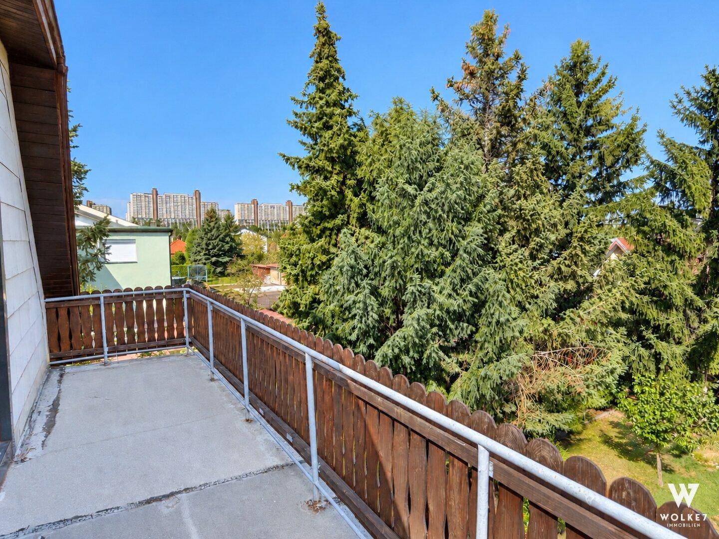 Geräumiger Balkon mit Holzzaun und weitem Blick ins Grüne unter strahlend blauem Himmel.