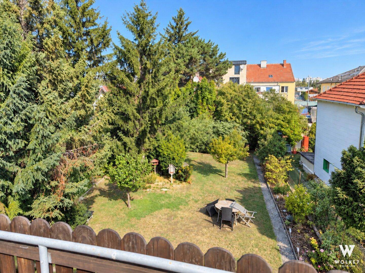 Blick von oben auf den gepflegten Garten mit Rasenfläche und Sitzgelegenheiten umgeben von Bäumen.