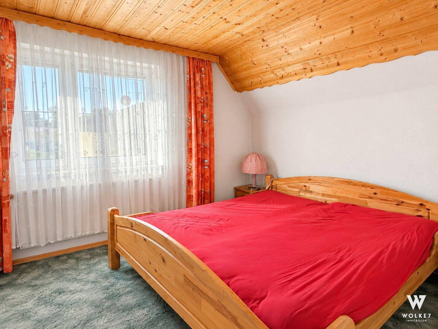 Gemütliches Schlafzimmer mit Holzbett, Teppichboden und Holzdecke unter einem großen Fenster.