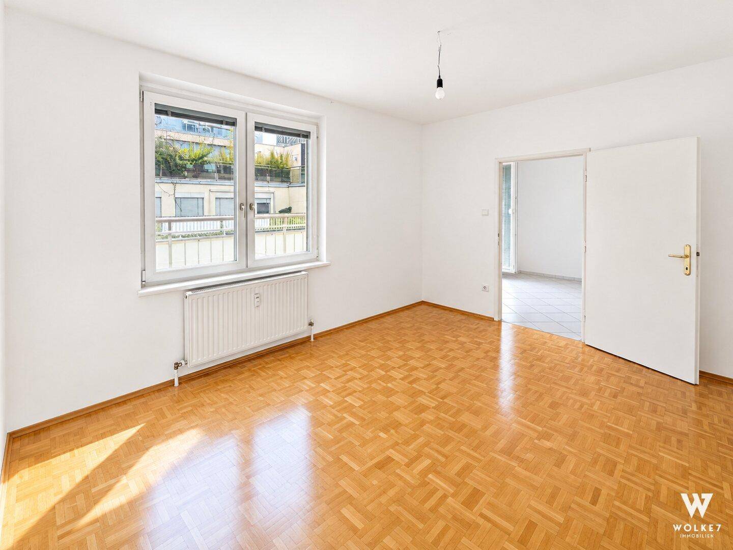 Helles Zimmer mit Parkettboden und großem Fenster mit Blick auf begrünte Dächer.