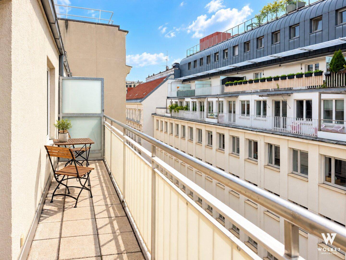 Schmaler Balkon mit Fliesenboden und kleinem Tisch und Stühlen, umgeben von Gebäuden.