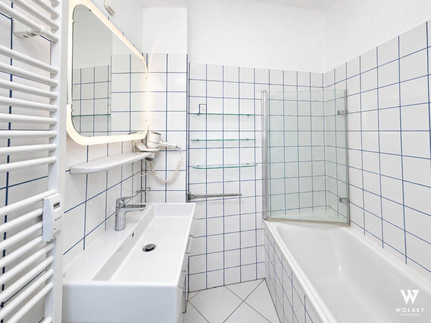 Helles Badezimmer mit weißen Fliesen, Badewanne mit Duschwand und einem modernen Waschbecken.
