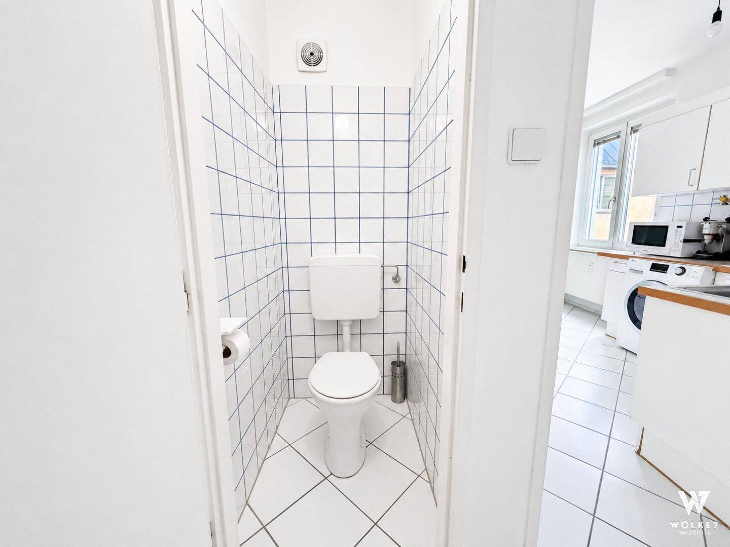 Separate Toilette mit weißen Fliesen und Lüftungsanlage, direkt neben der Küche gelegen.
