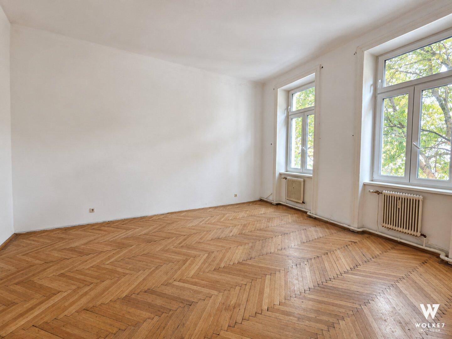 Geräumiges, leeres Zimmer mit Fischgrätparkettboden und großen Fenstern, die viel Licht hereinlassen.