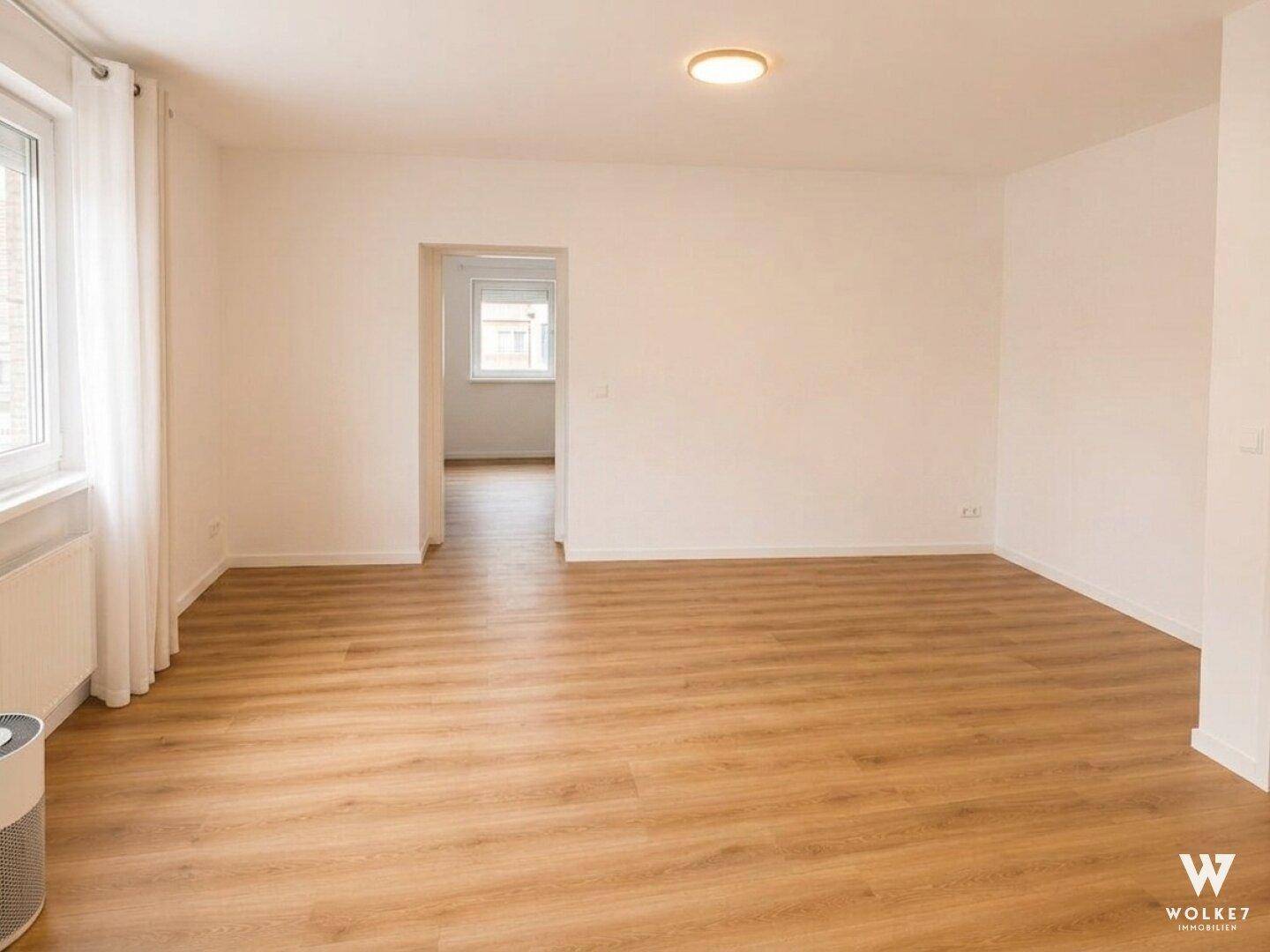 Geräumiger, leerer Wohnbereich mit hellem Holzboden und großen Fenstern.