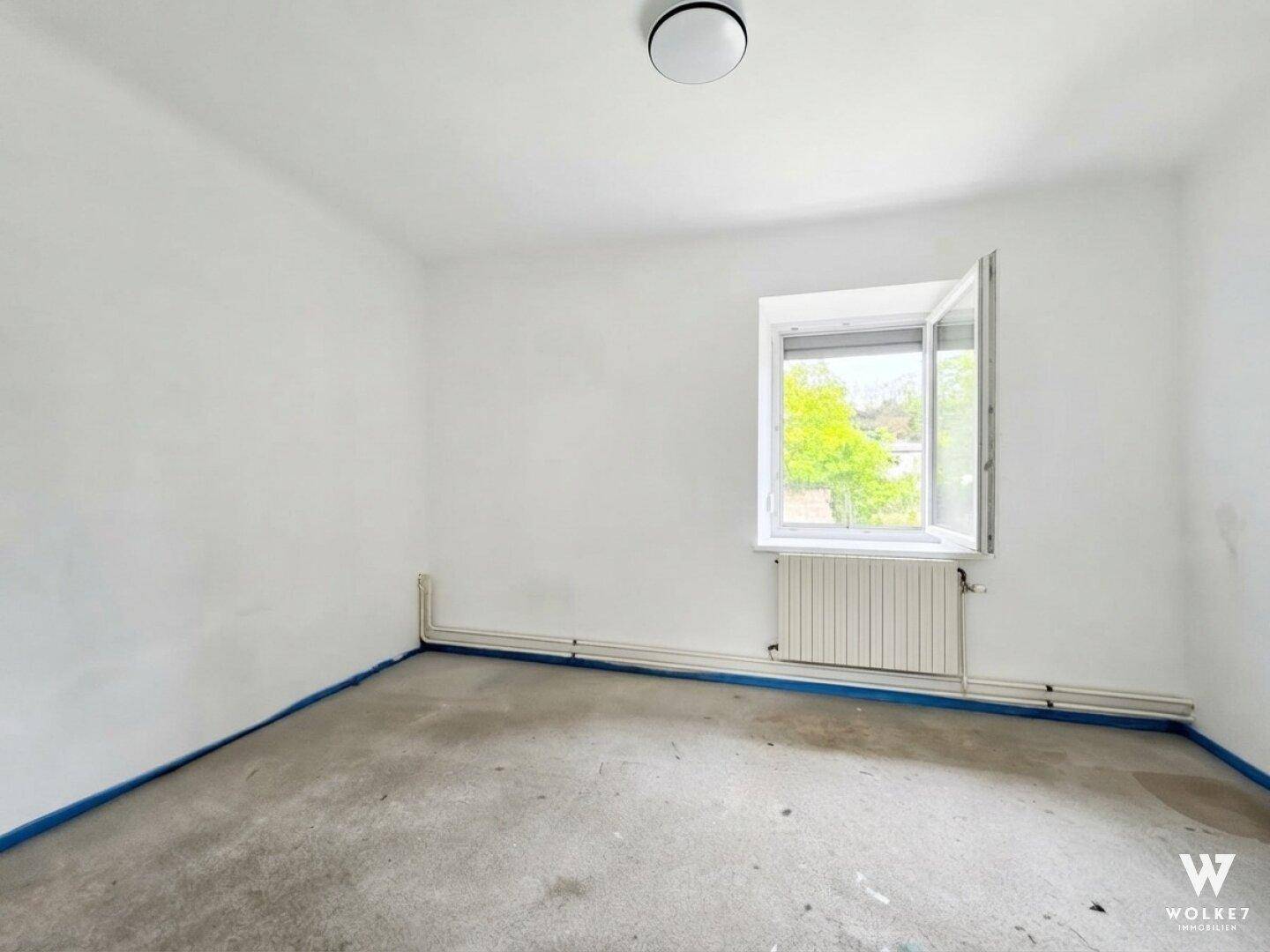 Helles, leeres Zimmer mit weiß gestrichenen Wänden, einem Fenster und Heizkörper.