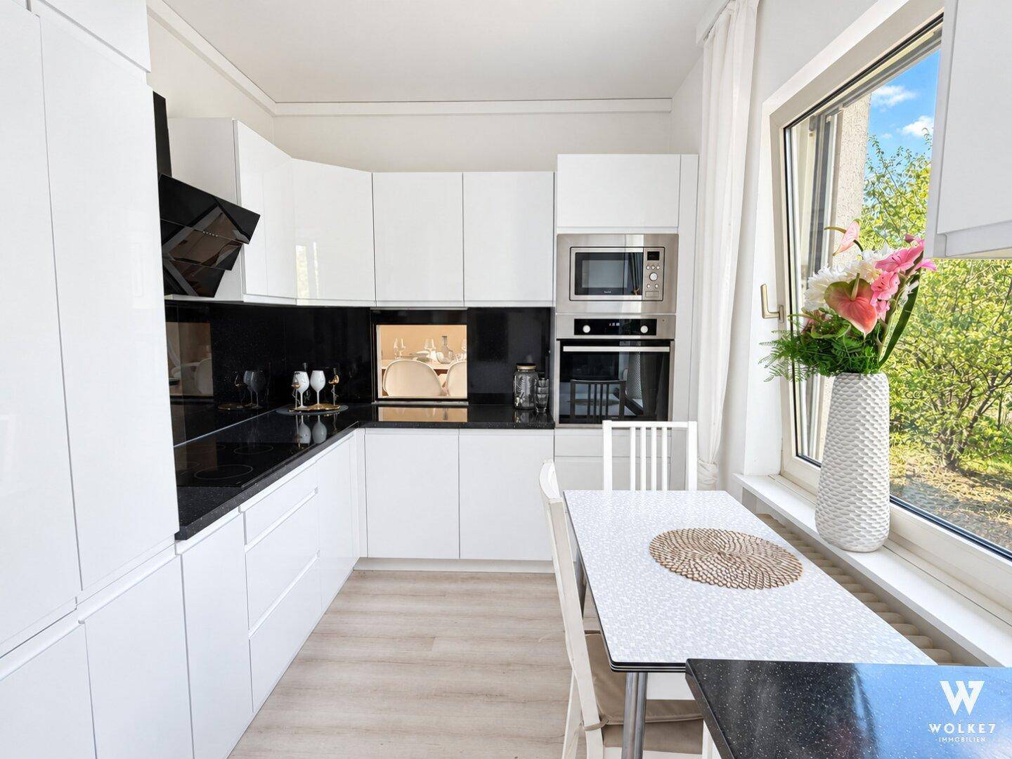 Moderne Küche mit weißen Hochglanzfronten, schwarzen Arbeitsplatten und Essbereich am Fenster.