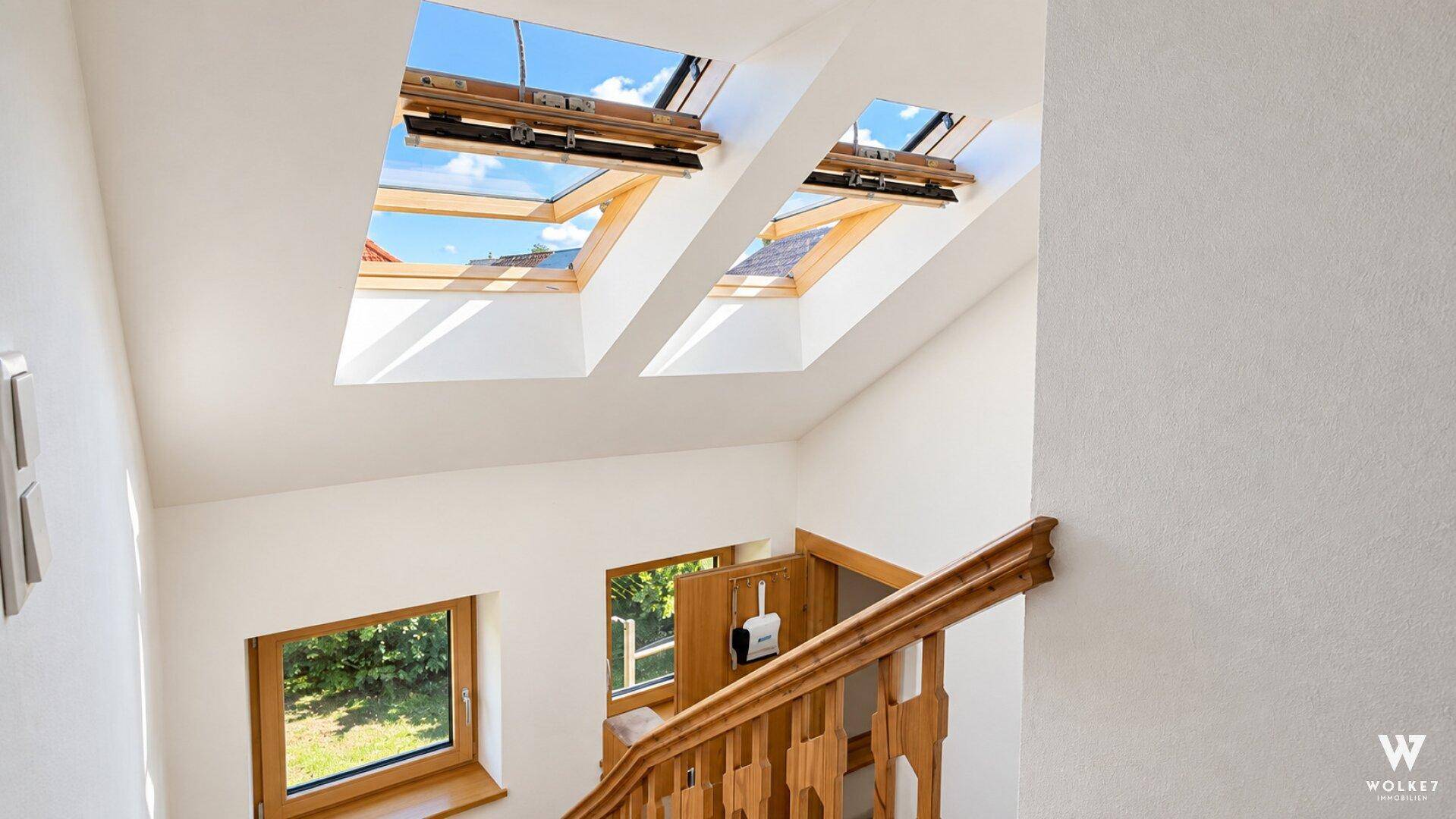 Helles Treppenhaus mit Holzgeländer und mehreren Dachfenstern, die viel Tageslicht hereinlassen.