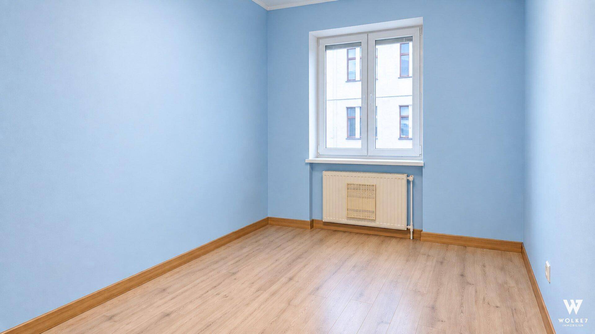 Leeres Zimmer mit blauen Wänden, Holzboden und einem Fenster, ideal als Kinder- oder Gästezimmer.