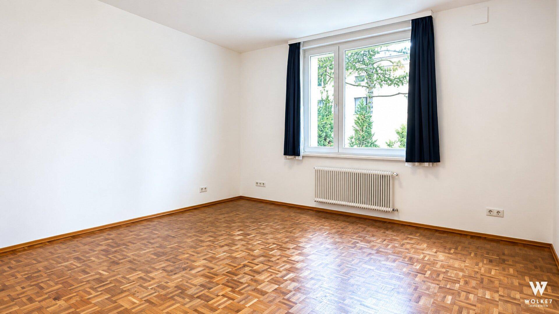 Heller Raum mit Parkettboden, weißen Wänden und einem Fenster mit Blick ins Grüne.