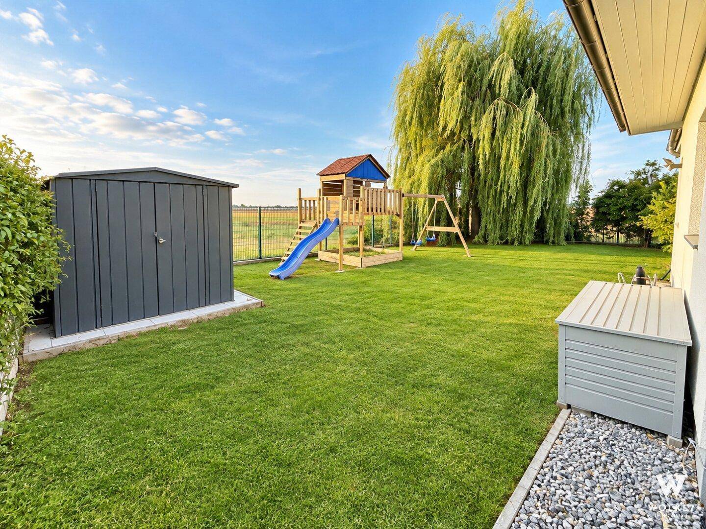 Großer Garten mit Rasenfläche, Spielplatz und Gartenhaus unter einem Weidenbaum.
