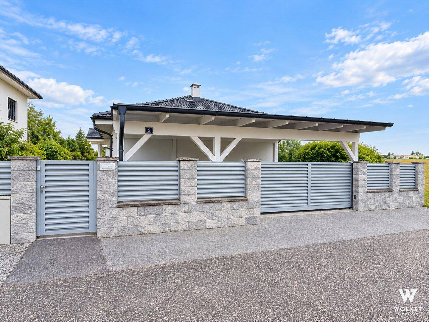 Moderne Hauszufahrt mit Zaun, Tor und Carport unter blauem Himmel.
