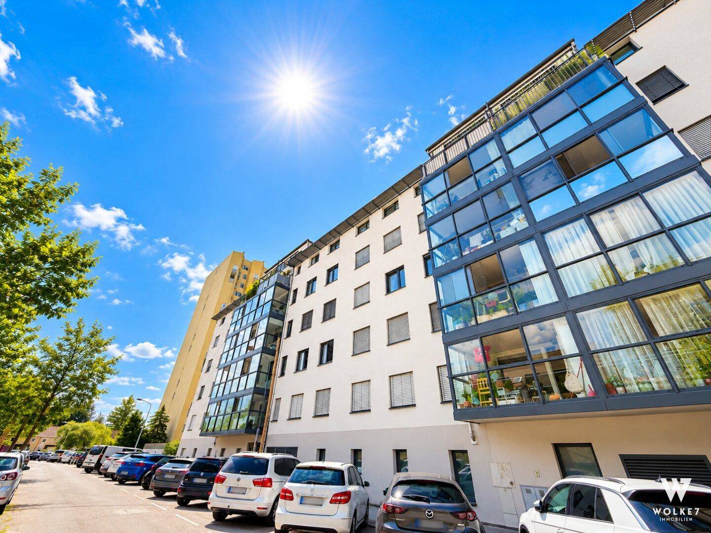 Moderne Wohnanlage mit Glasfassaden und Balkonen an einem sonnigen Tag, mit Parkplätzen davor.