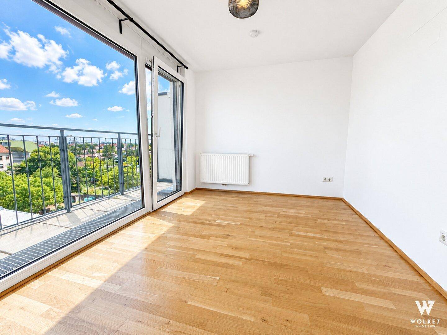 Heller, leerer Raum mit großem Fenster und Zugang zu einem Balkon, ausgestattet mit Holzfußboden.
