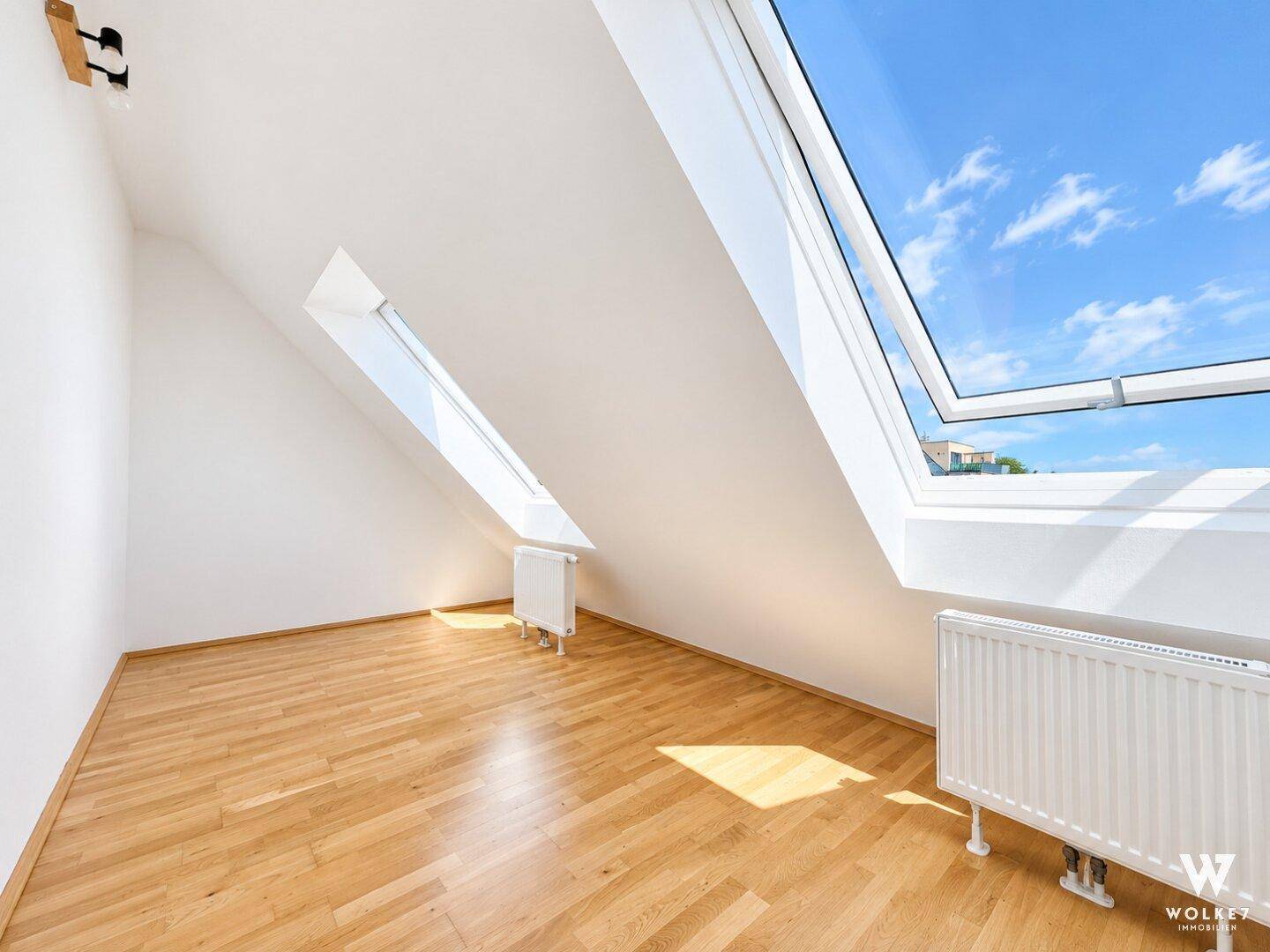 Leerer Raum im Dachgeschoss mit schräger Decke, großem Dachfenster und hellem Holzfußboden.