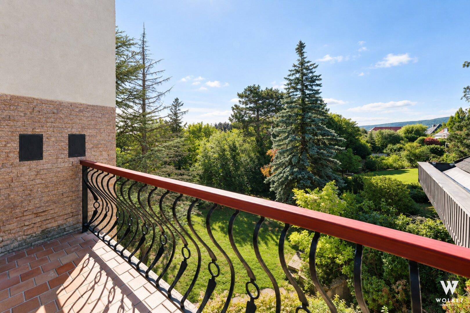 Blick von einem Balkon mit schmiedeeisernem Geländer auf einen üppigen grünen Garten und Bäume.