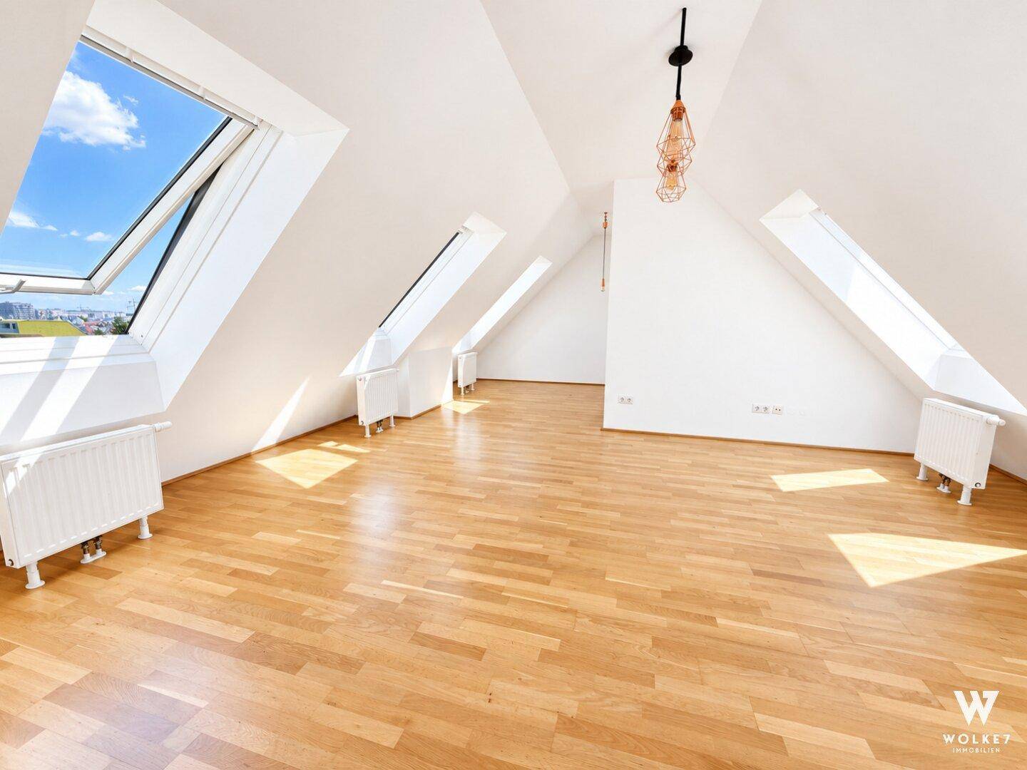 Heller, leerer Raum im Dachgeschoss mit schrägen Wänden, mehreren Dachfenstern und Holzfußboden.