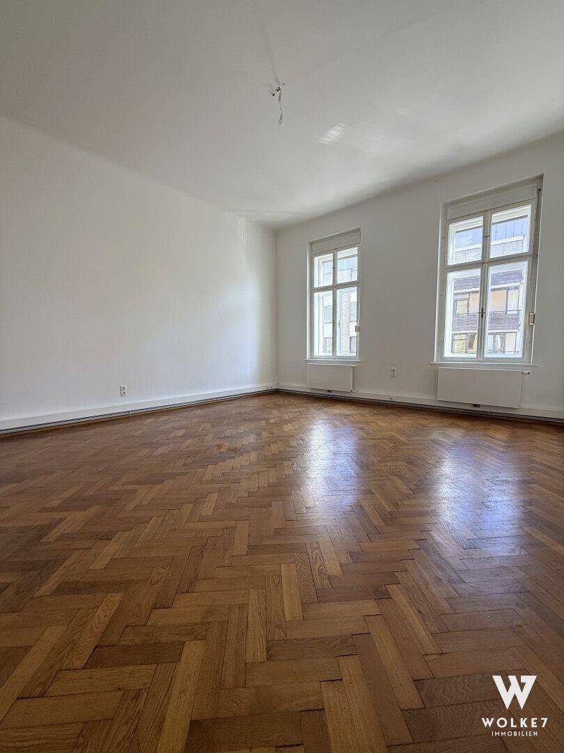 Lichtdurchflutetes Zimmer mit Fischgrätparkett und zwei großen Fenstern.