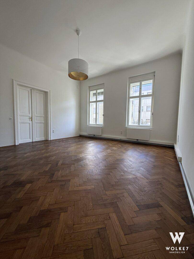 Geräumiges Zimmer mit Fischgrätparkett, großen Fenstern und einer modernen Hängelampe.