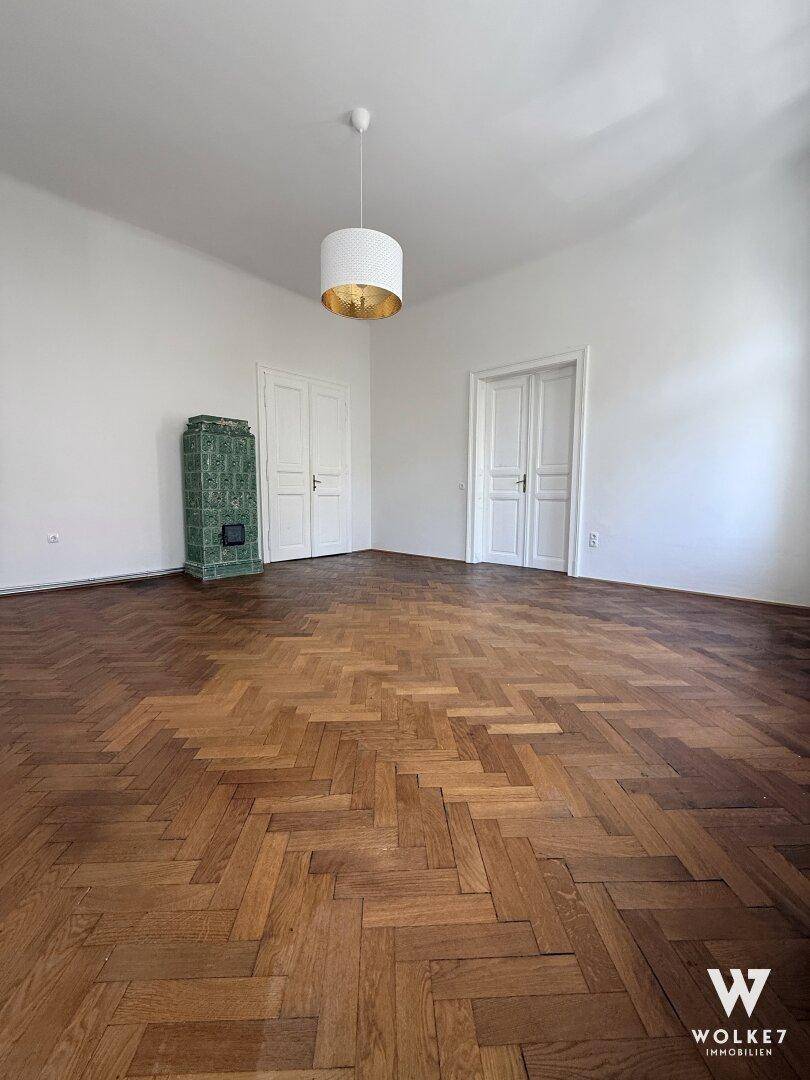 Helles Zimmer mit Fischgrätparkett und einem traditionellen grünen Kachelofen als Blickfang.
