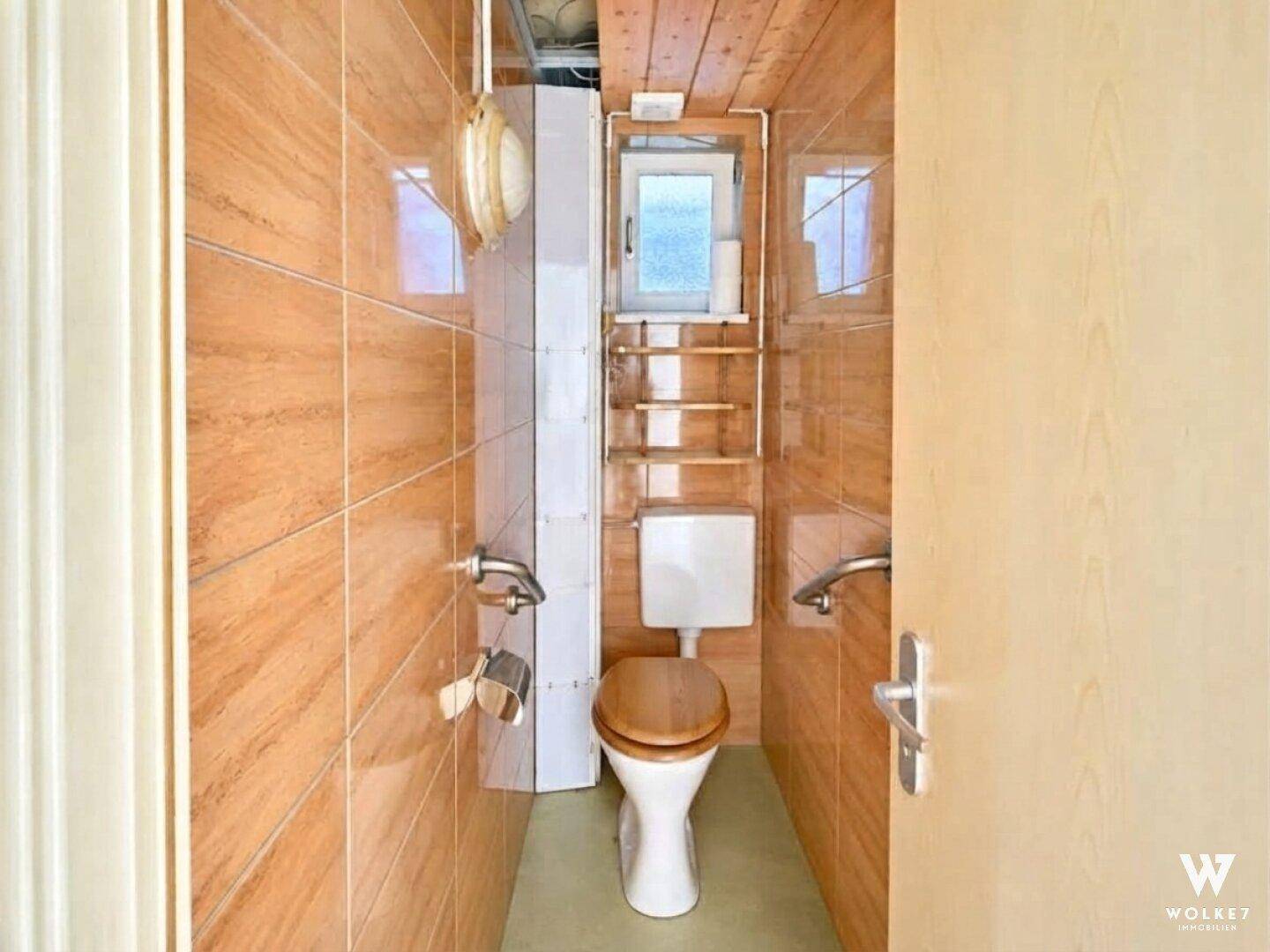 Kleines WC mit braunen Fliesen, Holzdecke, Fenster und Haltegriffen an der Wand.
