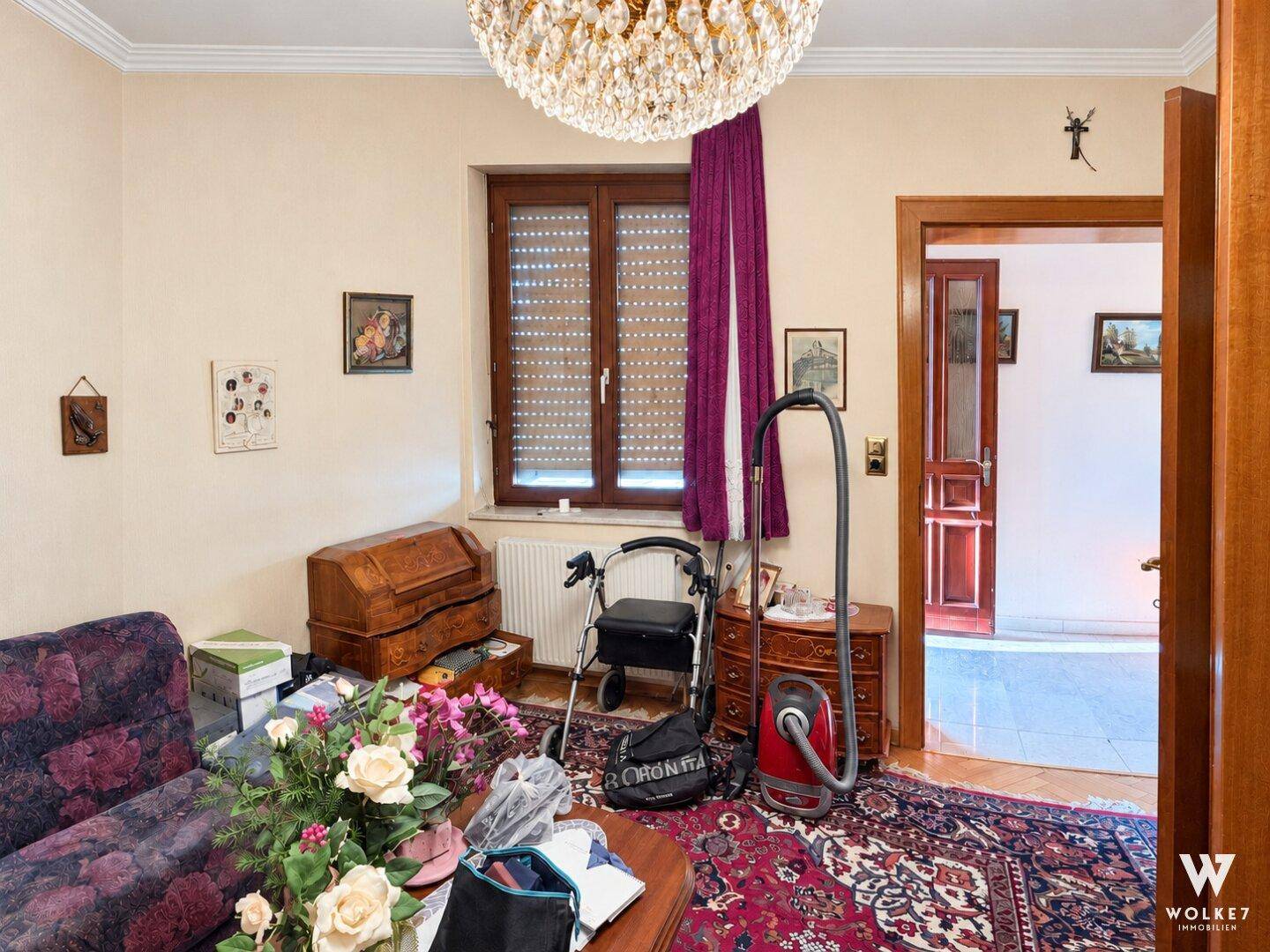 Wohnzimmer mit Teppichboden, Fenster mit Rollladen und verschiedenen Möbelstücken.