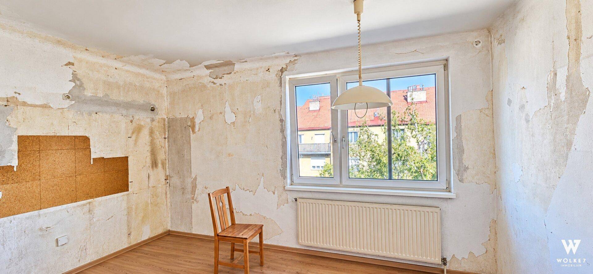 Unmöblierter Raum mit abgelöster Tapete, Parkettboden und einem Fenster mit Blick nach draußen.