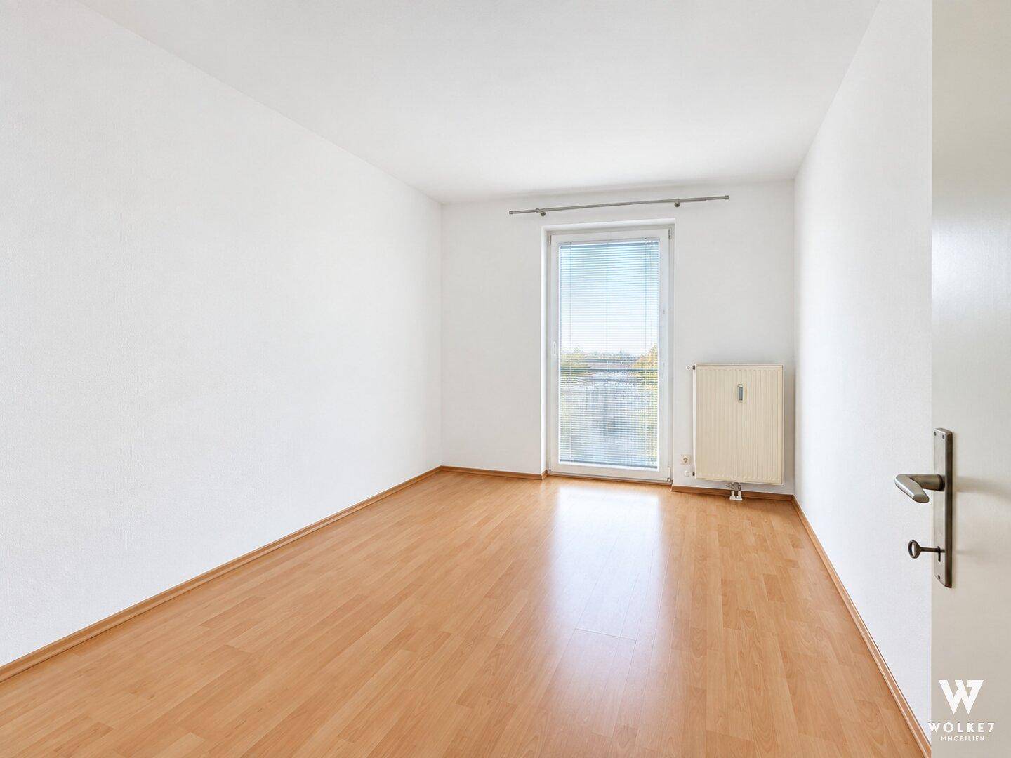 Helles, leeres Zimmer mit Laminatboden und Fenster mit Blick ins Grüne.