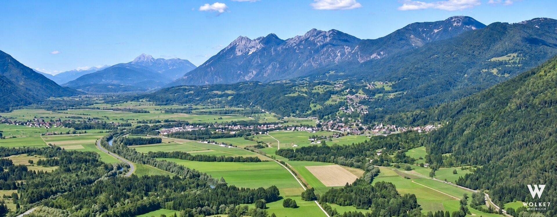 Panoramablick über ein weites Tal mit grünen Feldern, einer Siedlung und imposanten Bergen.