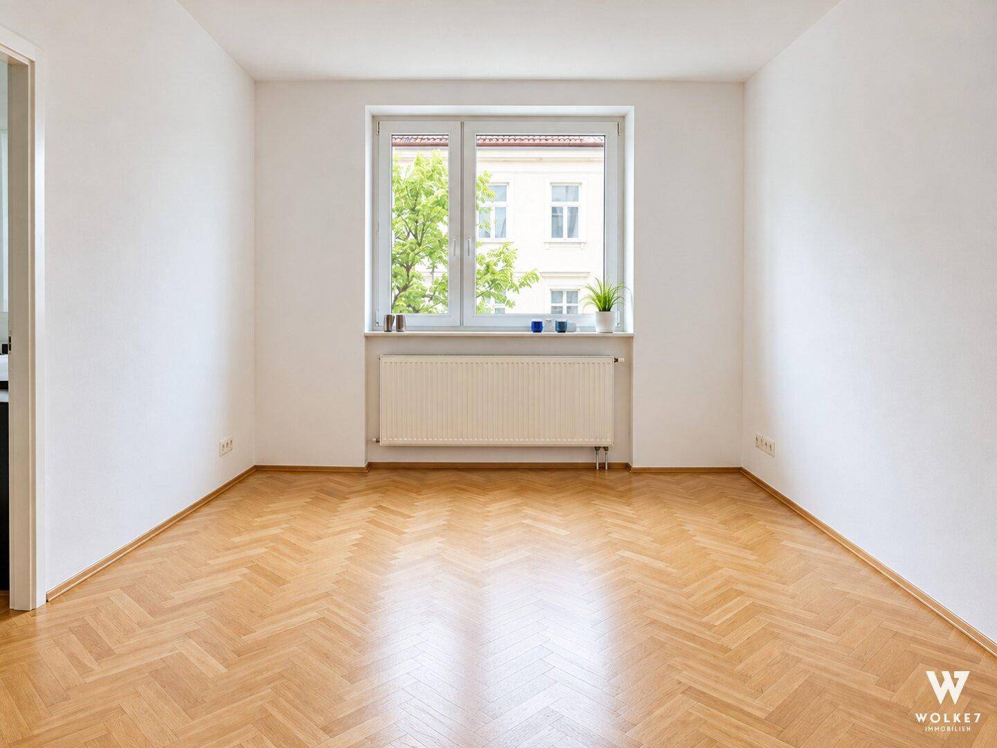 Leerer Raum mit hellem Fischgrätparkett, weißen Wänden und einem Fenster mit grüner Aussicht.