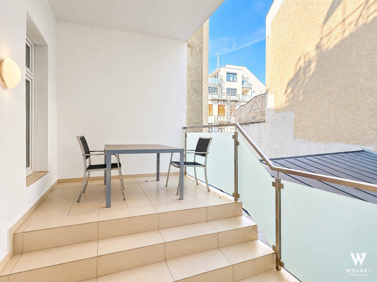Balkon mit Tisch und Stühlen, umgeben von Gebäuden unter blauem Himmel.
