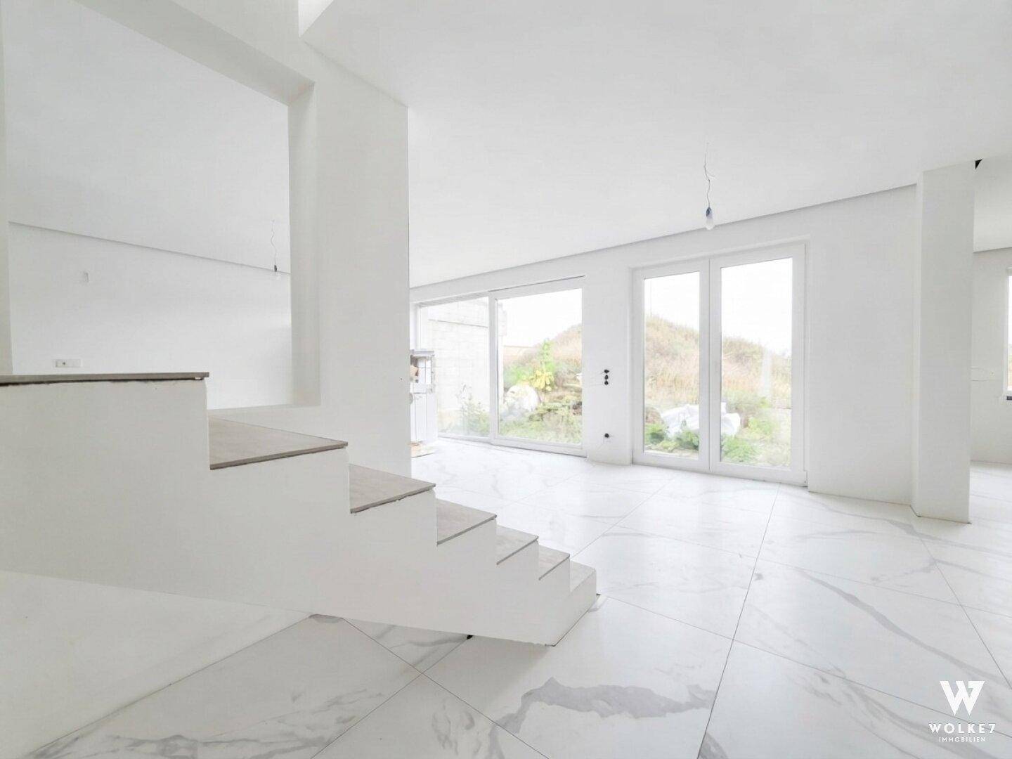 Geräumiger Wohnbereich im Neubau mit moderner Treppe, großen Fenstern und Fliesenboden.