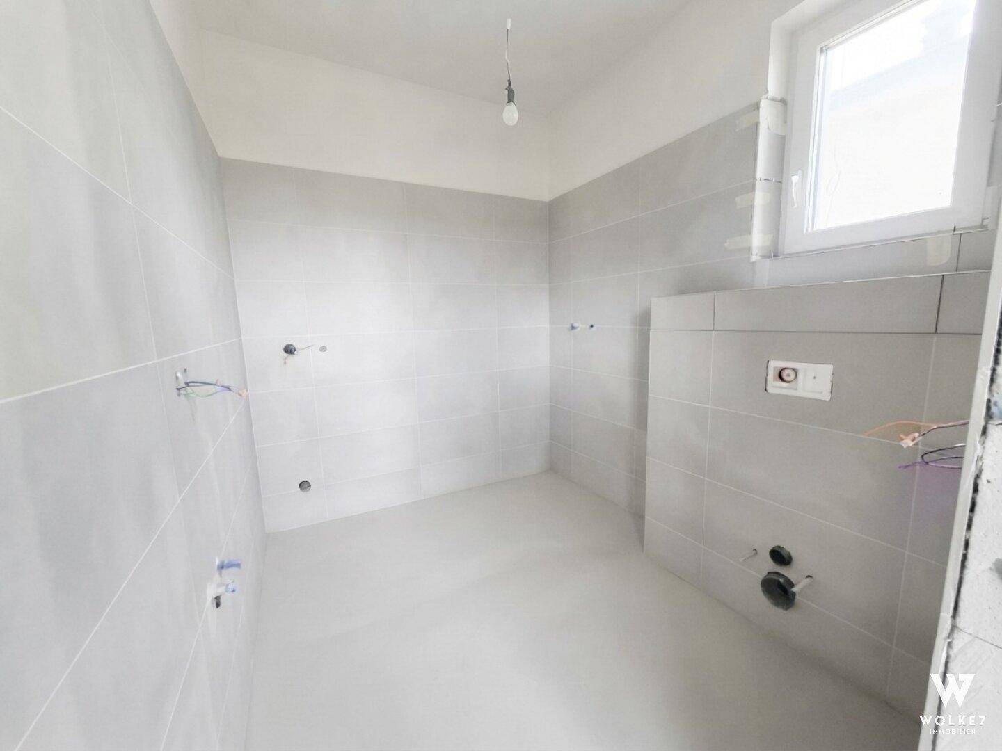 Modernes Badezimmer im Rohbau mit grauen Fliesen und Fenster für Tageslicht.