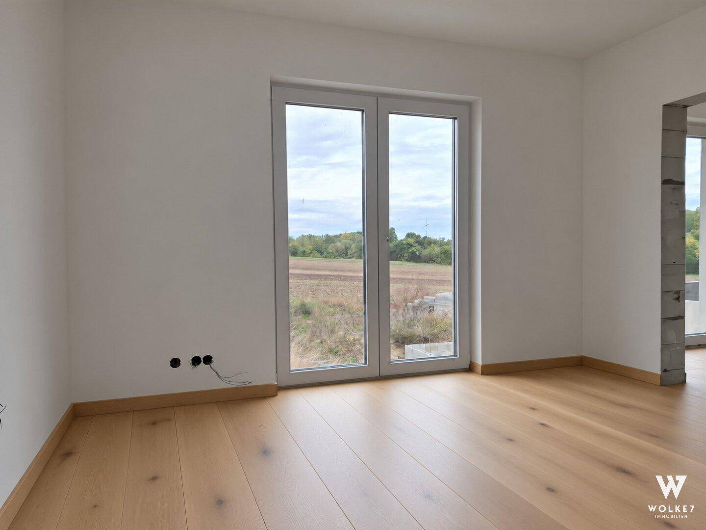 Heller Raum mit Holzboden und großen Fenstertüren zum Außenbereich.