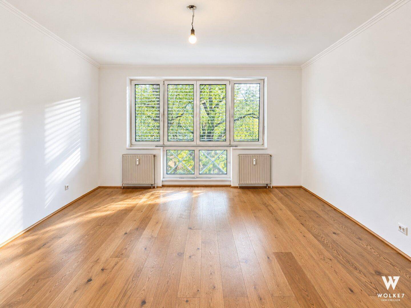 Geräumiges Wohnzimmer mit großen Fenstern, Holzboden und einem angenehmen Blick ins Grüne.