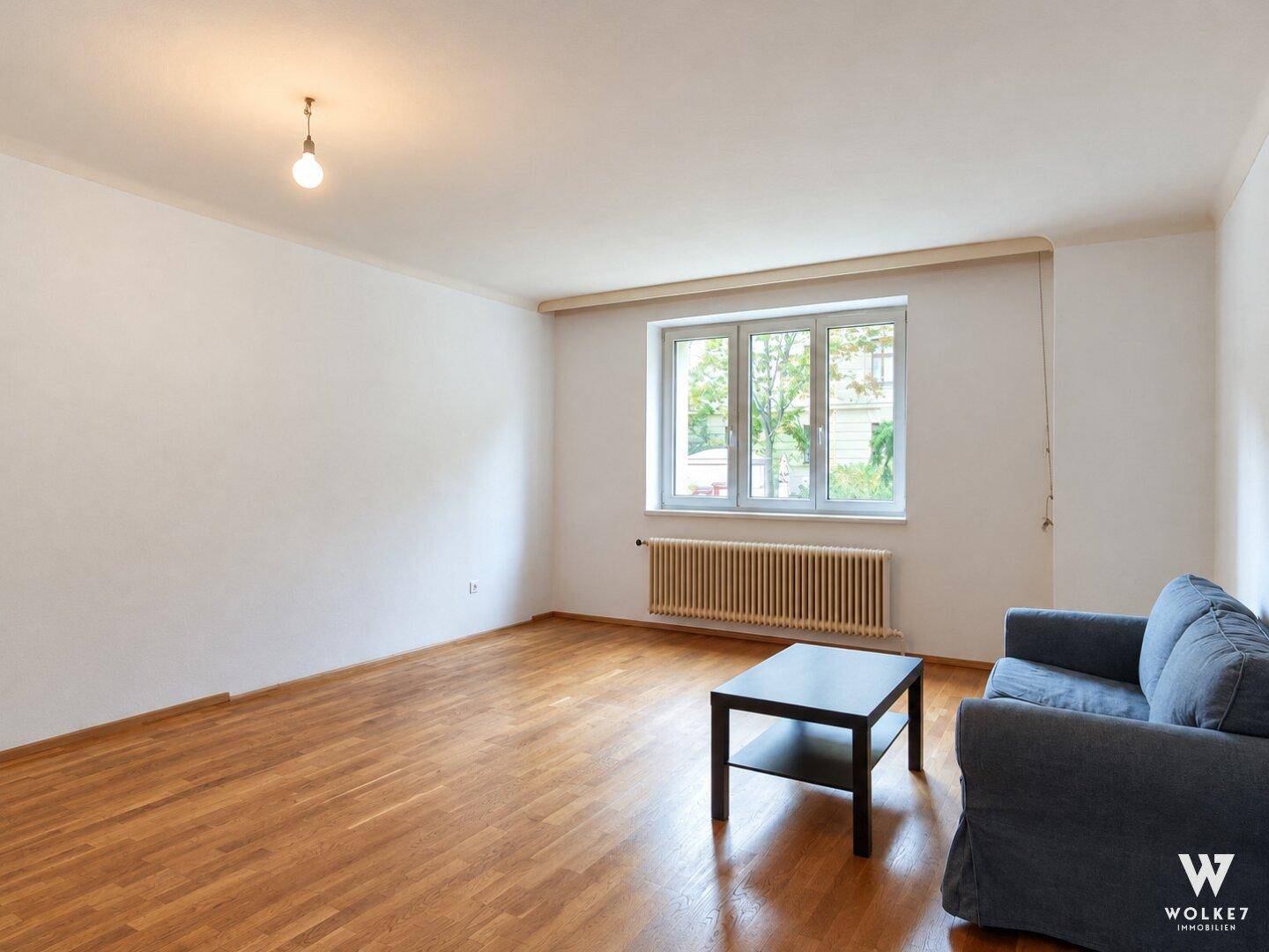 Geräumiges Wohnzimmer mit Holzboden, einem Fenster und einfacher Deckenbeleuchtung.
