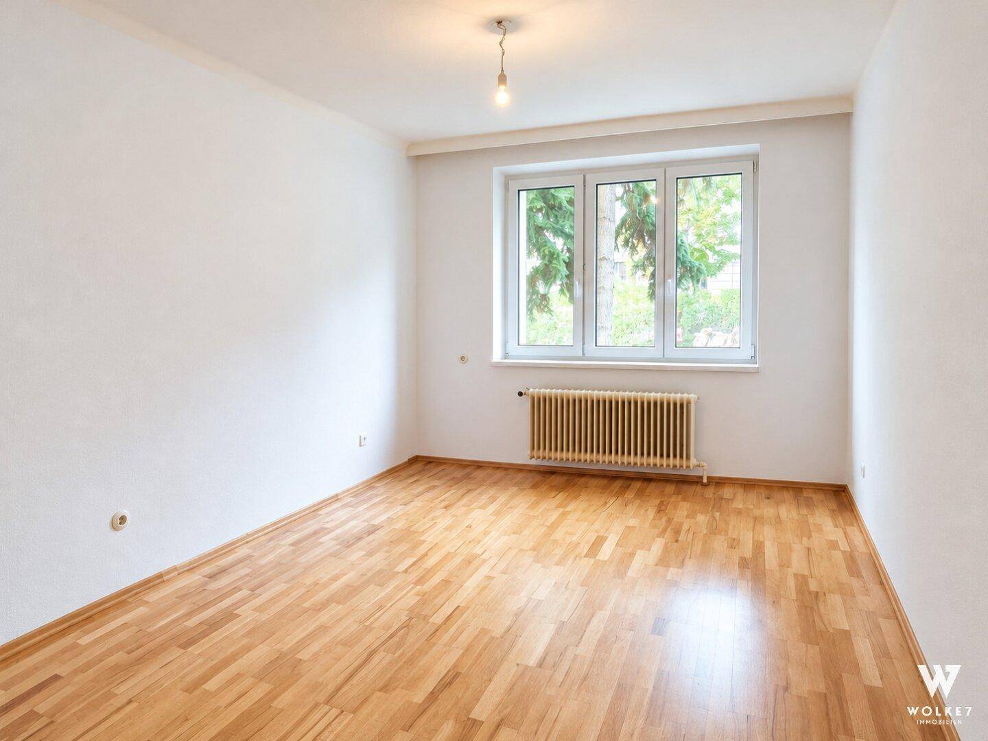 Helles, leeres Zimmer mit Holzboden und einem großen Fenster für viel Tageslicht.