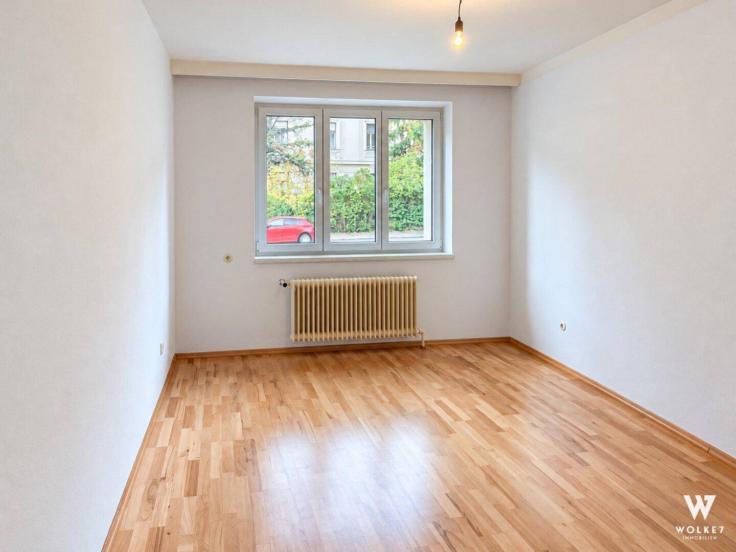 Geräumiges Zimmer mit Parkettboden, weißen Wänden und Blick auf die Straße.