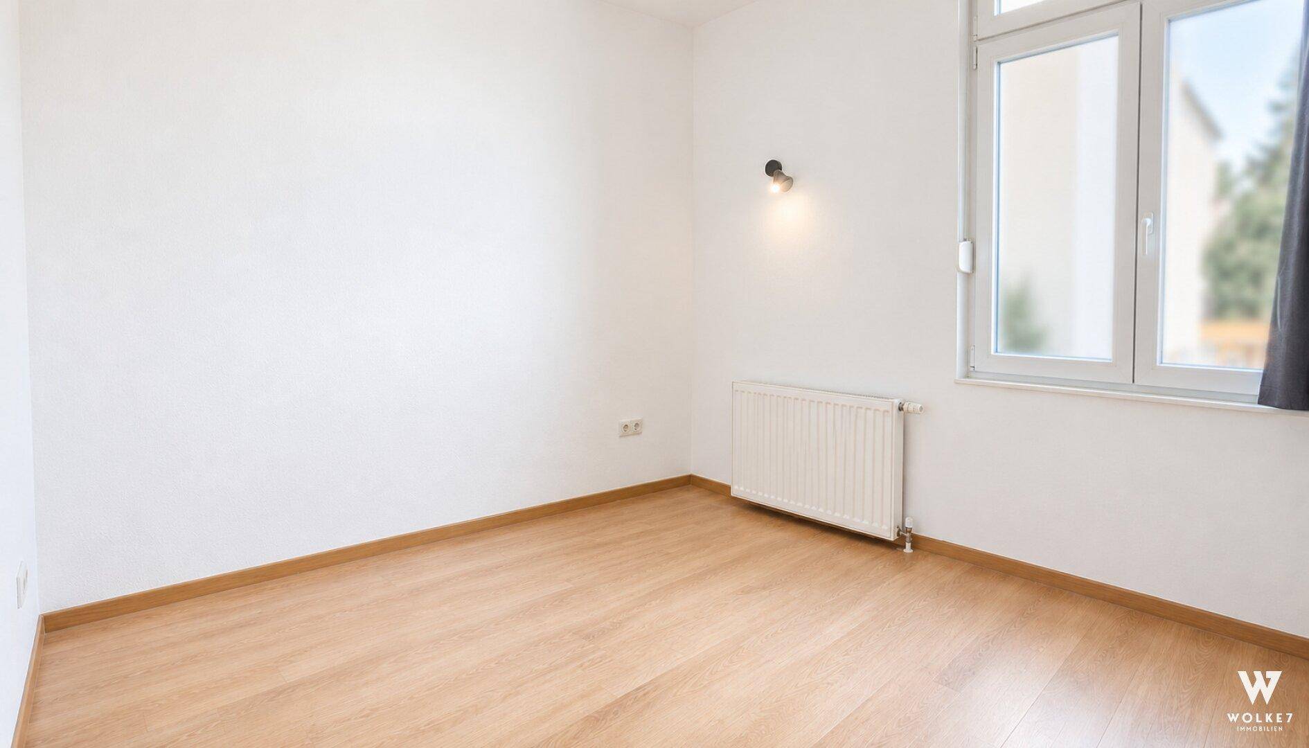 Heller Raum mit weiß gestrichenen Wänden, hellem Holzfußboden, großem Fenster und Heizkörper.