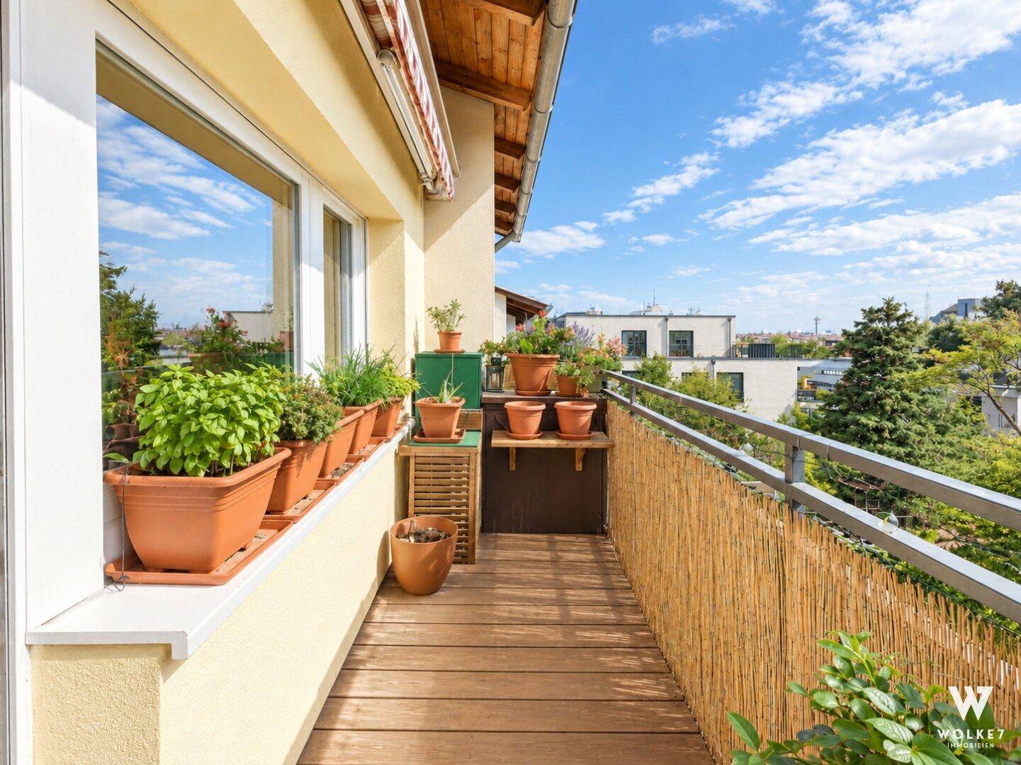 Sonniger Balkon mit Holzfußboden, vielen Pflanzen und Blick auf die Stadt und ins Grüne.