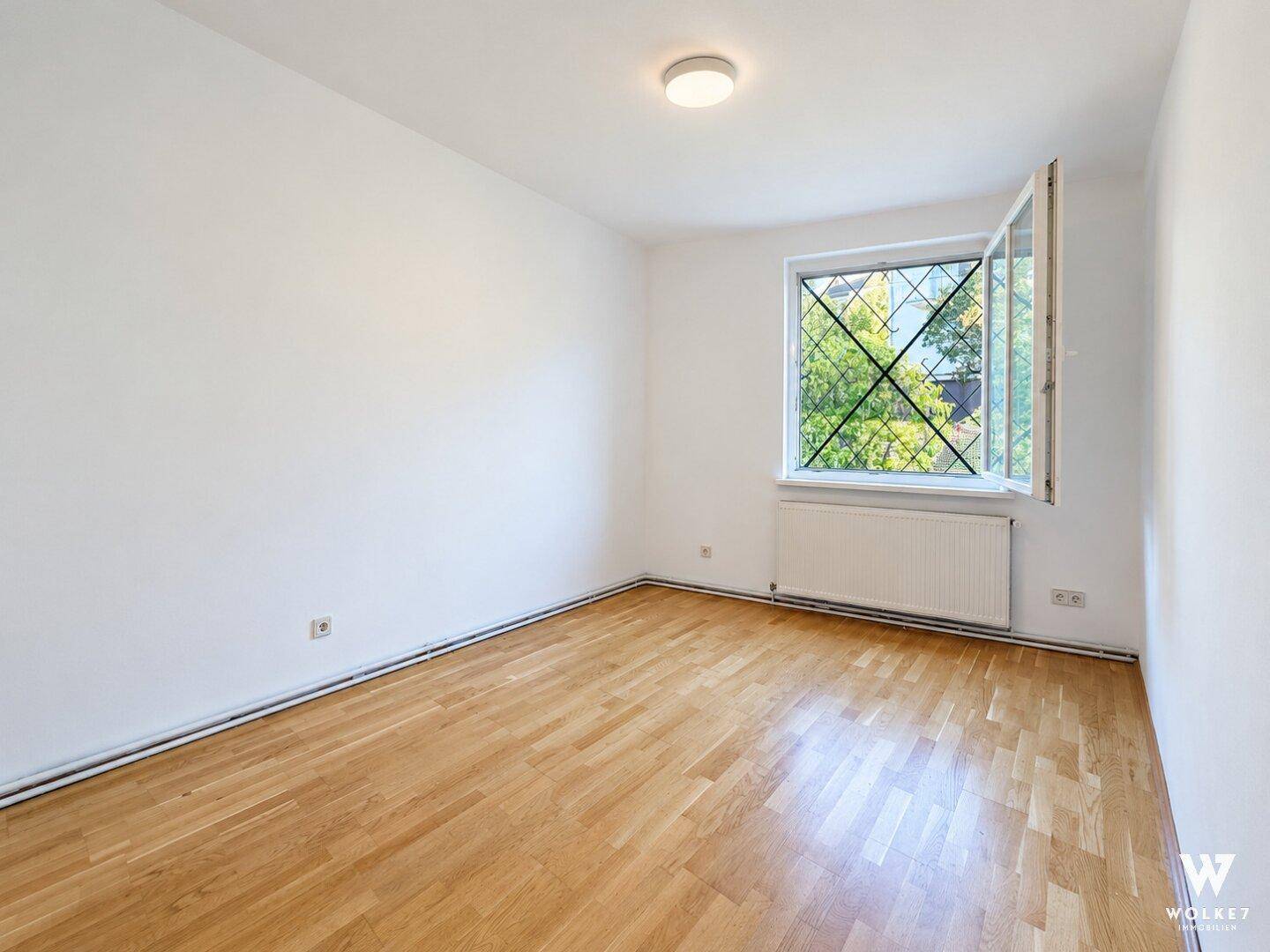 Leeres Zimmer mit hellem Parkettboden und einem Fenster, das viel Tageslicht hereinlässt.