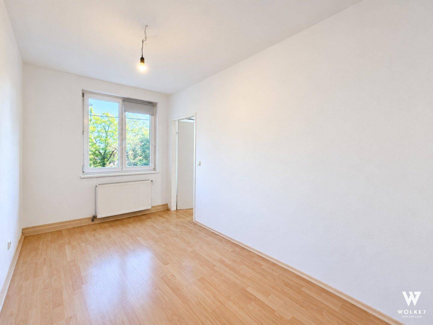 Freundliches Zimmer mit Parkettboden, weißen Wänden und einem Fenster mit Blick ins Grüne.