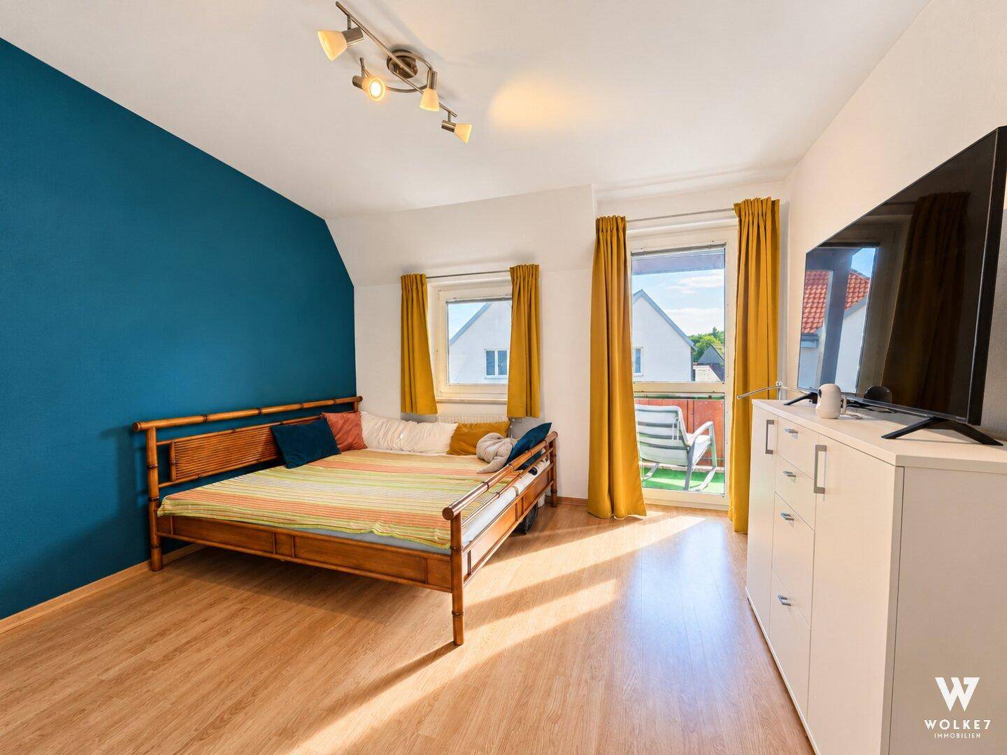 Schlafzimmer mit Holzbett, blauer Akzentwand und Zugang zu einem kleinen Balkon.