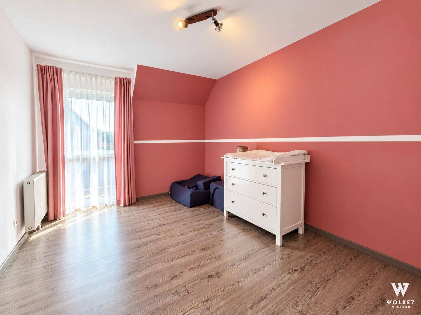 Geräumiges Kinderzimmer mit rosa Akzentwand, Wickelkommode und großem Fenster.