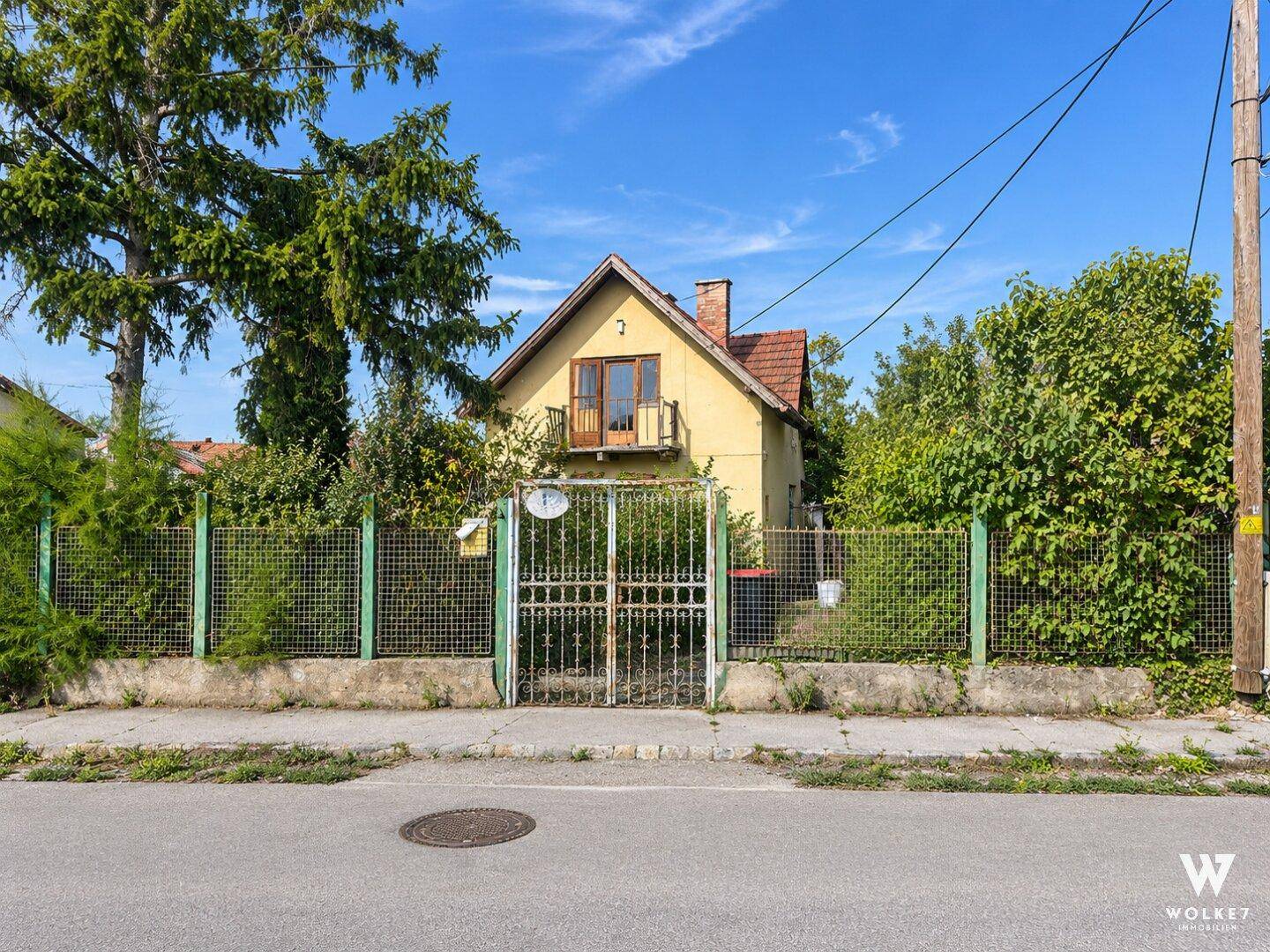 Älteres gelbes Einfamilienhaus mit kleinem Balkon und Zaun, von der Straße aus gesehen.