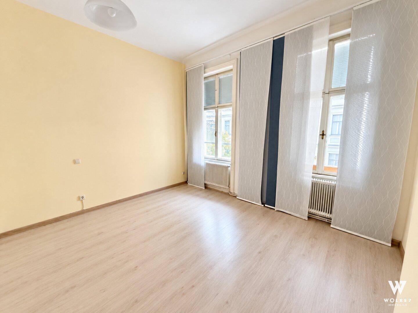 Geräumiges Zimmer mit hellgelben Wänden, Laminatboden und großen Fenstern für natürliche Belichtung.
