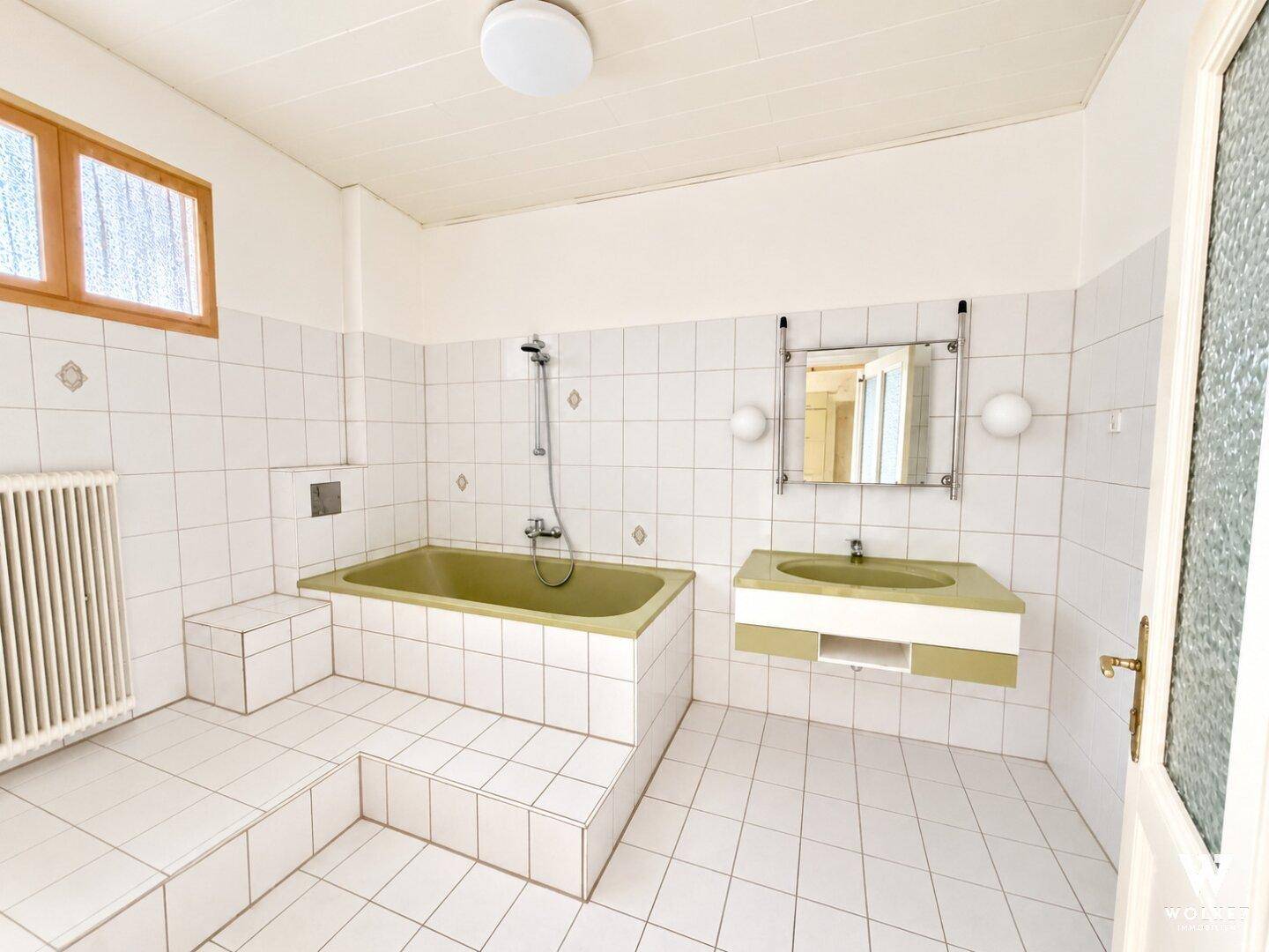 Badezimmer mit weißen Fliesen, einer Badewanne in Olivgrün und einem Fenster für natürliche Belüftung.