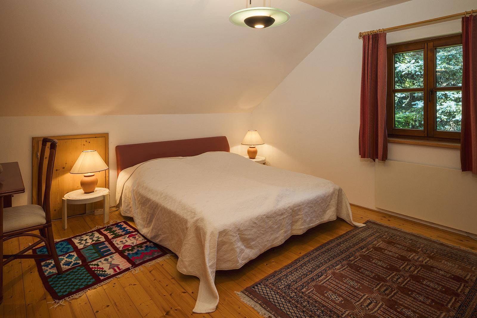 Schlafzimmer im Dachgeschoss mit schrägen Wänden, Teppichen und Blick ins Grüne.