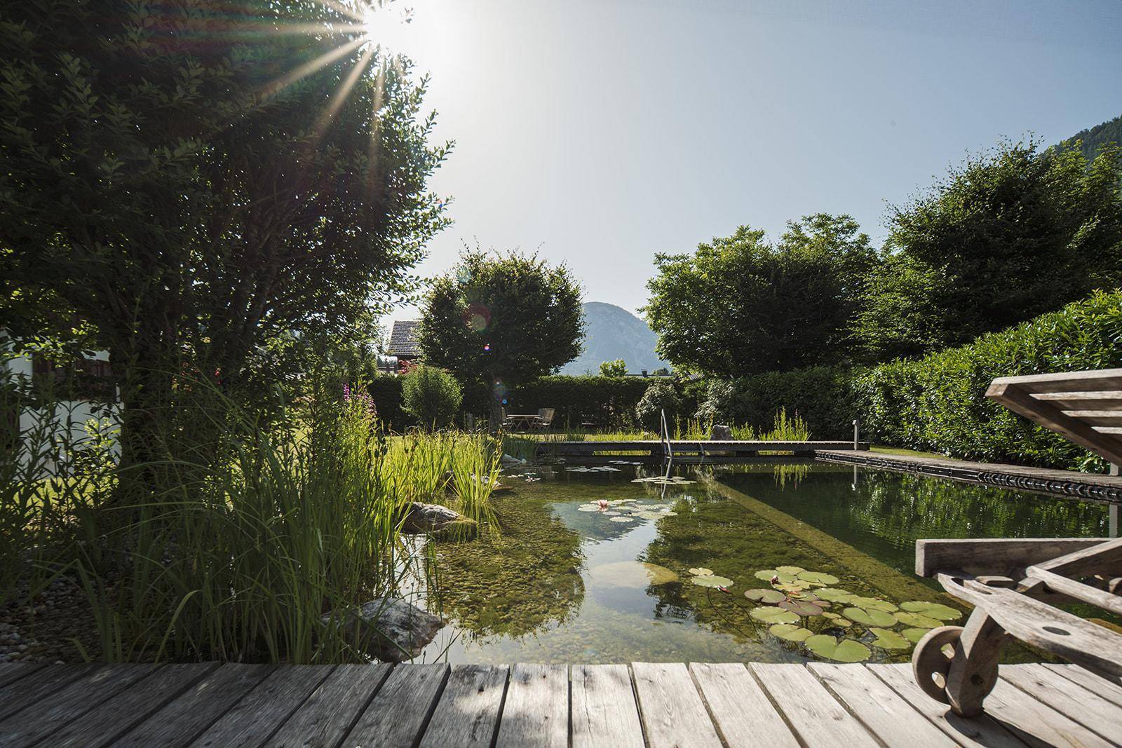 Idyllischer Naturpool mit Holzdeck und Liegestuhl, umgeben von üppigem Grün und Bergblick.