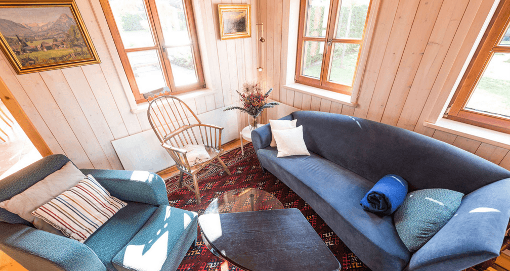 Kleiner, heller Wohnbereich mit blauer Couch, Holzwänden und Fenstern mit Blick ins Grüne.